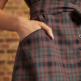 Close-up of a dress's front pocket with black contrast piping, showing a hand tucked inside and the brown plaid fabric with turquoise windowpane stripes.