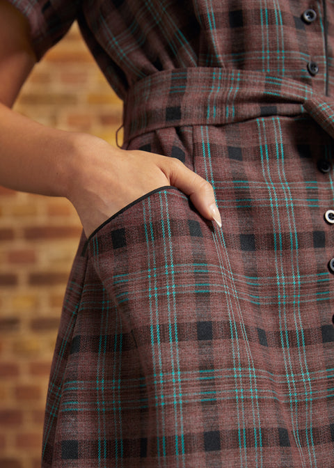 Close-up of a dress's front pocket with black contrast piping, showing a hand tucked inside and the brown plaid fabric with turquoise windowpane stripes.