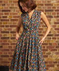 A woman wearing a sleeveless, knee-length vintage inspired dress with a colorful mushroom print on a dark background, standing against a brick wall.