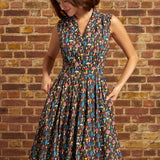 A woman wearing a sleeveless, knee-length vintage inspired dress with a colorful mushroom print on a dark background, standing against a brick wall.