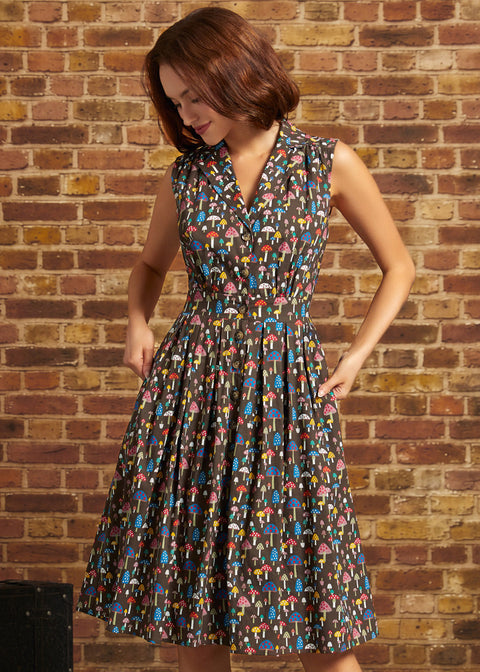 A woman wearing a sleeveless, knee-length vintage inspired dress with a colorful mushroom print on a dark background, standing against a brick wall.
