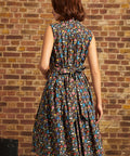 A woman wearing a sleeveless, knee-length A-line dress with a colorful abstract pattern on a dark background, photographed from the back against a brick wall.
