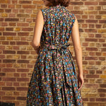 A woman wearing a sleeveless, knee-length A-line dress with a colorful abstract pattern on a dark background, photographed from the back against a brick wall.