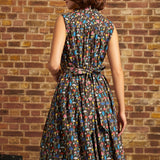 A woman wearing a sleeveless, knee-length A-line dress with a colorful abstract pattern on a dark background, photographed from the back against a brick wall.