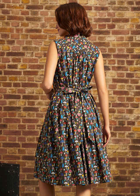 A woman wearing a sleeveless, knee-length A-line dress with a colorful abstract pattern on a dark background, photographed from the back against a brick wall.