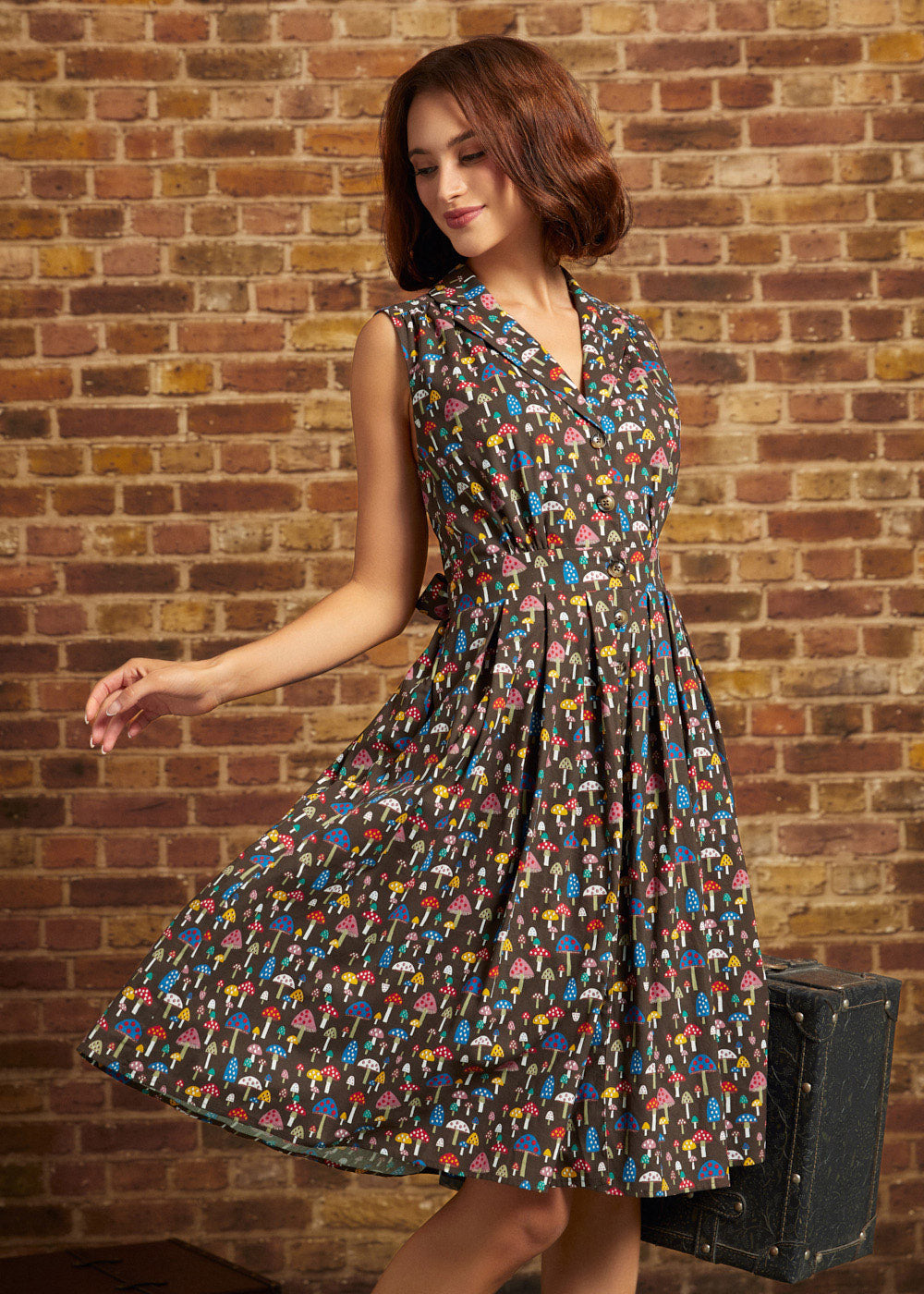 Model wearing the Carousel Cutie Dress in Mushroom Medley, a brown sleeveless cotton dress covered in colorful mushrooms, with a button-front bodice and flared skirt in motion against a brick wall.