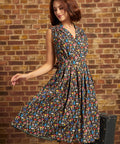Model wearing the Carousel Cutie Dress in Mushroom Medley, a brown sleeveless cotton dress covered in colorful mushrooms, with a button-front bodice and flared skirt in motion against a brick wall.