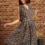 Model wearing the Carousel Cutie Dress in Mushroom Medley, a brown sleeveless cotton dress covered in colorful mushrooms, with a button-front bodice and flared skirt in motion against a brick wall.