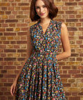 A woman wearing a sleeveless shirt dress with a colorful mushroom print on a dark background, standing in front of a brick wall.