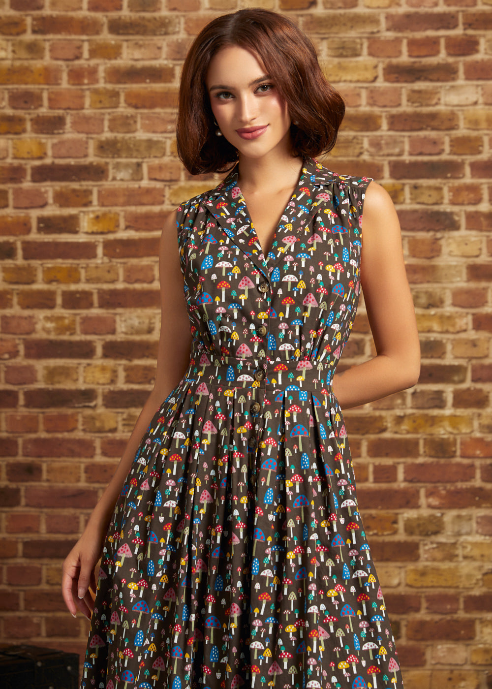 A woman wearing a sleeveless shirt dress with a colorful mushroom print on a dark background, standing in front of a brick wall.