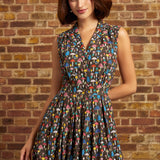A woman wearing a sleeveless shirt dress with a colorful mushroom print on a dark background, standing in front of a brick wall.