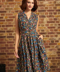 A woman wearing a sleeveless, knee-length A-line dress with a multicolor mushroom print on a dark background, standing against a rustic brick wall.