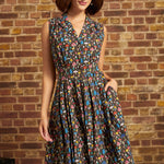 A woman wearing a sleeveless, knee-length A-line dress with a multicolor mushroom print on a dark background, standing against a rustic brick wall.