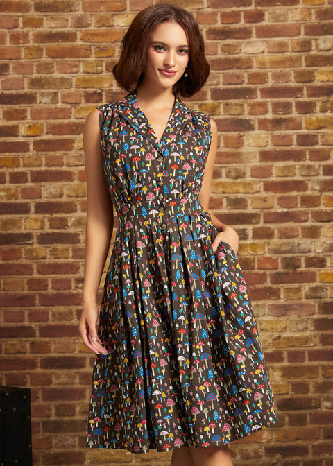 A woman wearing a sleeveless, knee-length A-line dress with a multicolor mushroom print on a dark background, standing against a rustic brick wall.