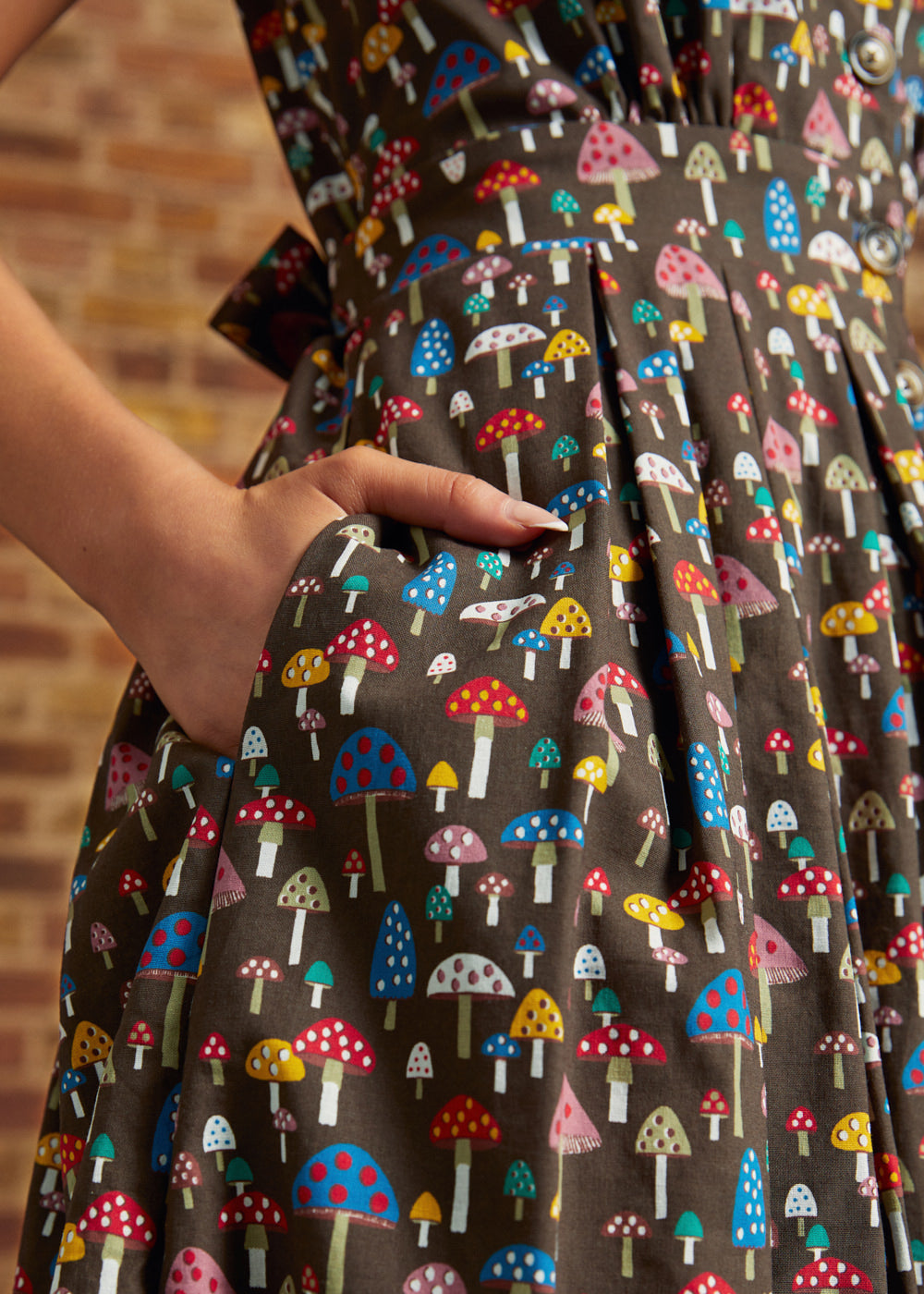 Close-up of a hand tucked into the side seam pocket of the Carousel Cutie Dress, showing colorful red, blue, pink, yellow, and white mushrooms against a dark brown background.