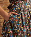 Close-up of a hand tucked into the side seam pocket of the Carousel Cutie Dress, showing colorful red, blue, pink, yellow, and white mushrooms against a dark brown background.