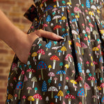 Close-up of a hand tucked into the side seam pocket of the Carousel Cutie Dress, showing colorful red, blue, pink, yellow, and white mushrooms against a dark brown background.