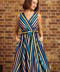 A woman wearing a sleeveless wrap-style midi dress with vertical multicolor stripes in blue, yellow, orange, purple, and black, standing against a brick wall.