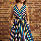 A woman wearing a sleeveless wrap-style midi dress with vertical multicolor stripes in blue, yellow, orange, purple, and black, standing against a brick wall.