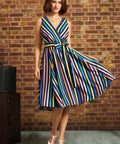 Model wearing ModCloth's Instant Energy Faux-Wrap Dress in Electric Avenue, featuring colorful vertical stripes, a V-neck bodice, tie waist, and flared skirt, paired with blue-and-white heels.