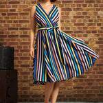 Model wearing ModCloth's Instant Energy Faux-Wrap Dress in Electric Avenue, featuring colorful vertical stripes, a V-neck bodice, tie waist, and flared skirt, paired with blue-and-white heels.
