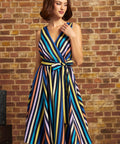 A woman wearing a sleeveless wrap-style midi dress with vertical multicolor stripes in blue, yellow, orange, pink, and white, standing against a brick wall.