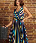 A woman models a sleeveless wrap-style midi dress with vertical multicolor stripes in blue, yellow, orange, purple, and black, standing against a rustic brick wall next to a vintage suitcase.