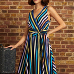 A woman models a sleeveless wrap-style midi dress with vertical multicolor stripes in blue, yellow, orange, purple, and black, standing against a rustic brick wall next to a vintage suitcase.