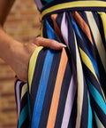 Close-up of model's hand tucked into the side pocket of the multicolored striped dress, showing blue, turquoise, yellow, peach, lavender, and cream stripes.