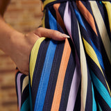 Close-up of model's hand tucked into the side pocket of the multicolored striped dress, showing blue, turquoise, yellow, peach, lavender, and cream stripes.