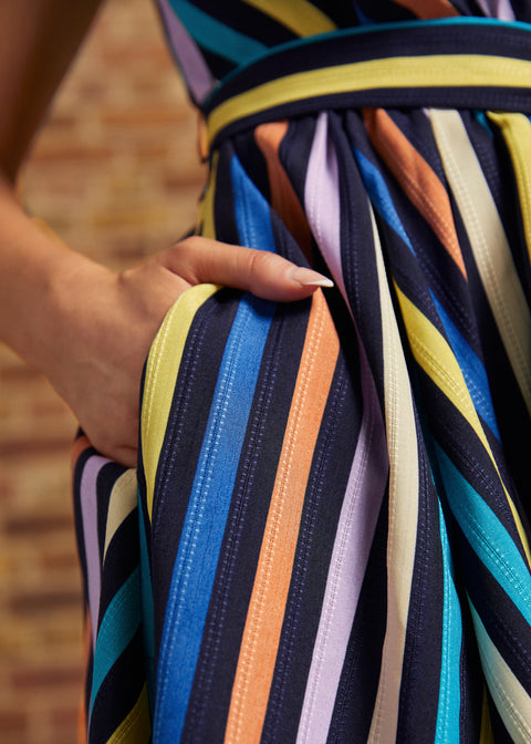 Close-up of model's hand tucked into the side pocket of the multicolored striped dress, showing blue, turquoise, yellow, peach, lavender, and cream stripes.