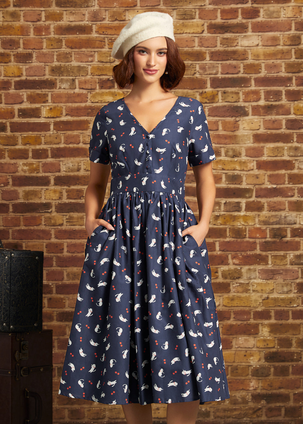 A woman wearing a navy blue short-sleeve midi dress with a white and red cat pattern, paired with a cream beret, standing against a rustic brick wall.