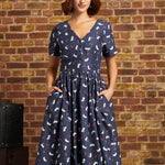 A woman wearing a navy blue short-sleeve midi dress with a white and red cat pattern, paired with a cream beret, standing against a rustic brick wall.