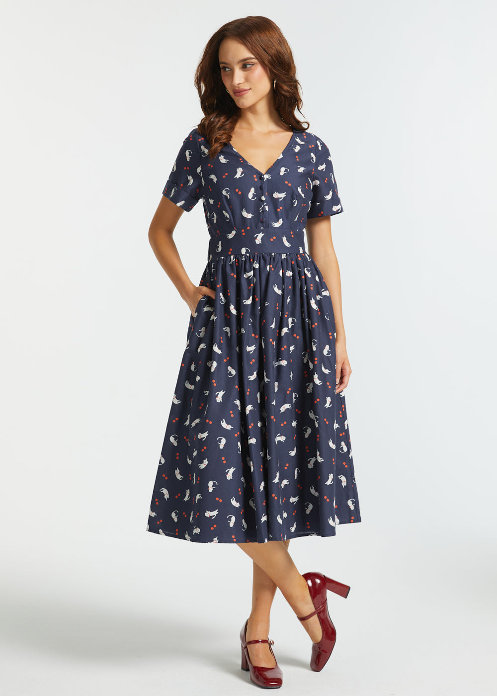 A woman modeling a navy blue midi dress with a white and red cat pattern, featuring short sleeves, a V-neckline, and a gathered skirt, paired with dark red heels against a light gray studio background.