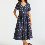 A woman modeling a navy blue midi dress with a white and red cat pattern, featuring short sleeves, a V-neckline, and a gathered skirt, paired with dark red heels against a light gray studio background.