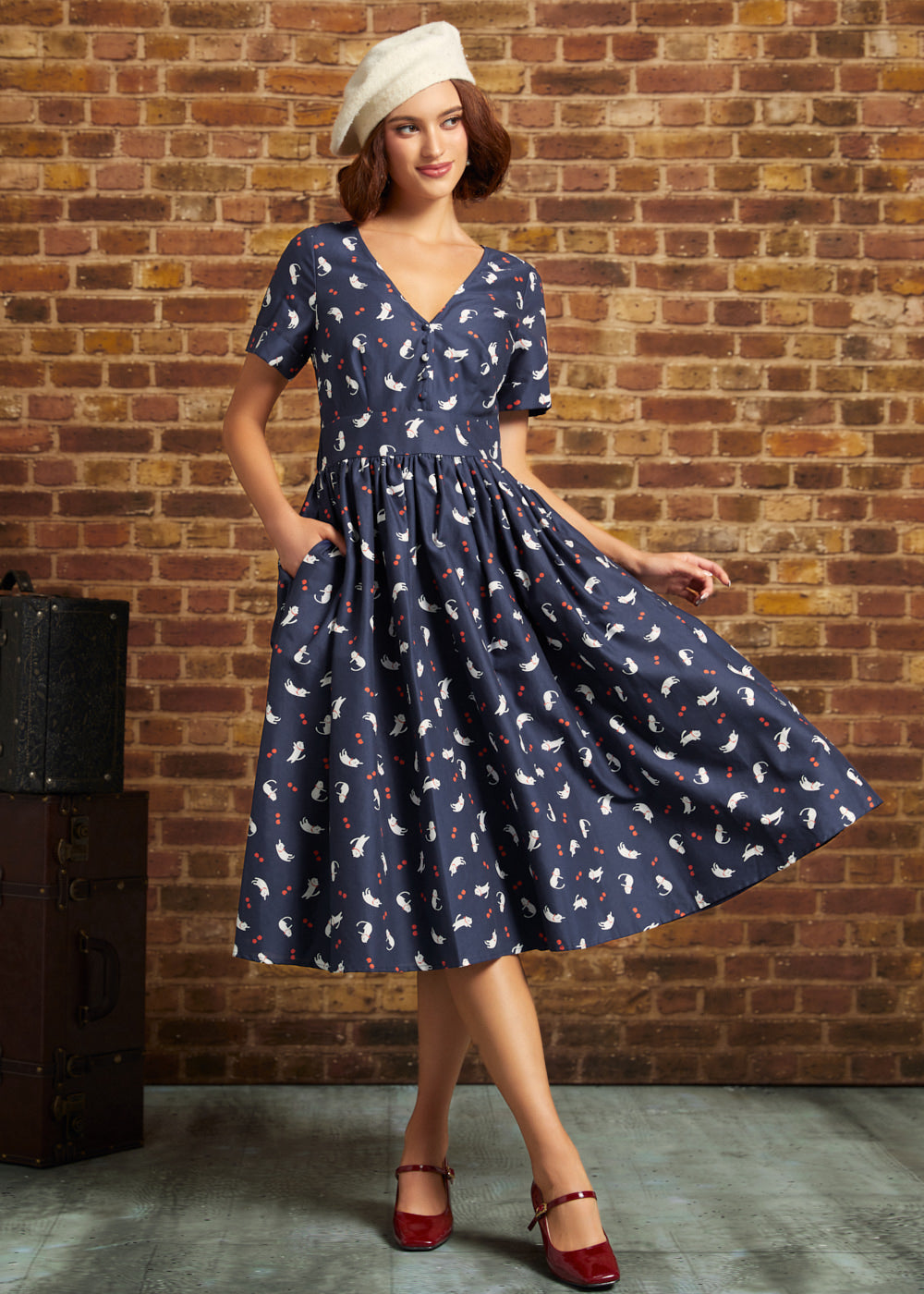 Model wearing a navy cat and berry print cotton midi dress with a fitted waist and flared skirt, styled with burgundy Mary Jane heels.