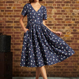 Model wearing a navy cat and berry print cotton midi dress with a fitted waist and flared skirt, styled with burgundy Mary Jane heels.