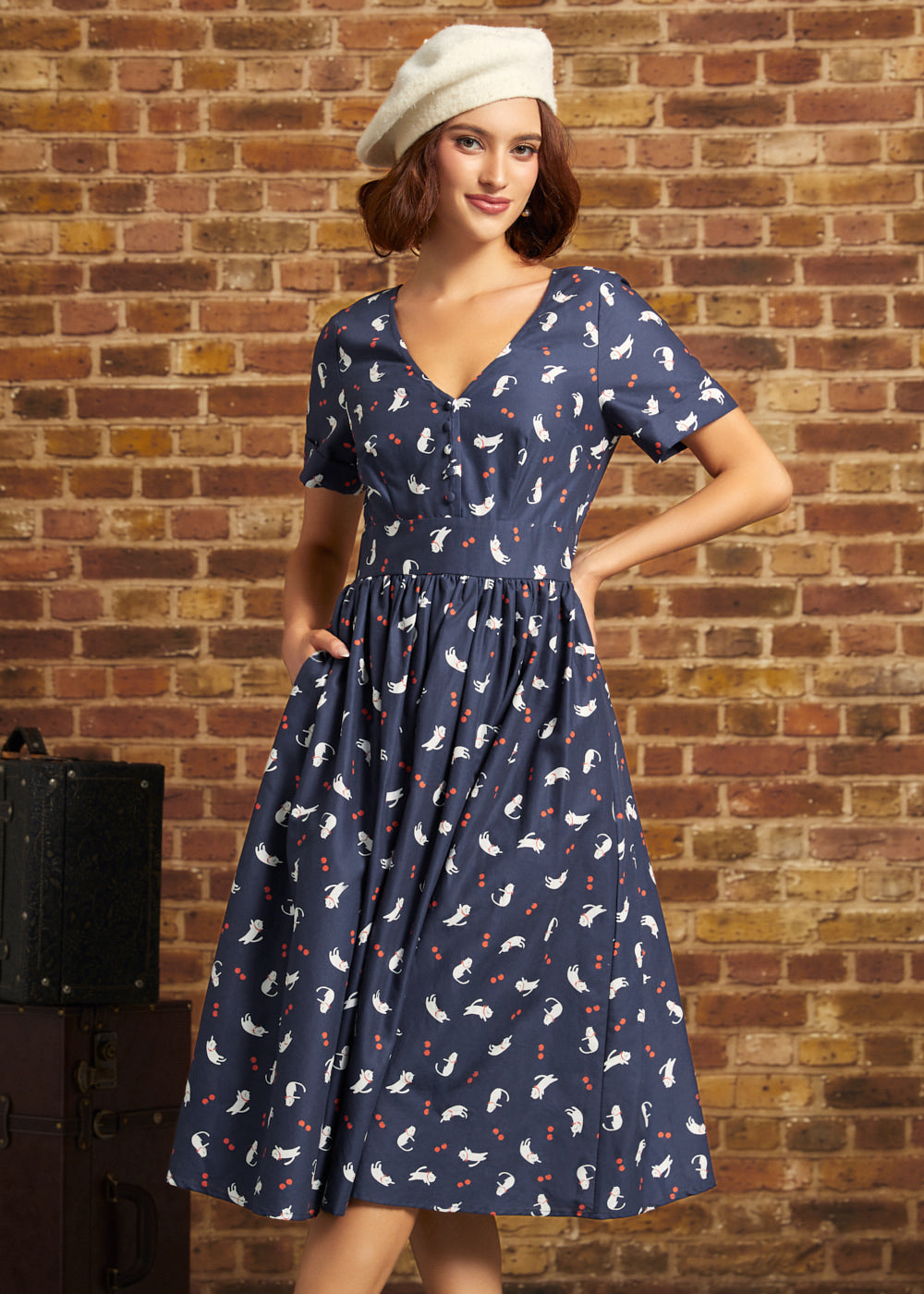 Model wearing a navy cotton fit-and-flare midi dress with white cat and red berry print, V-neckline, short sleeves, and side pockets, styled with a cream beret.