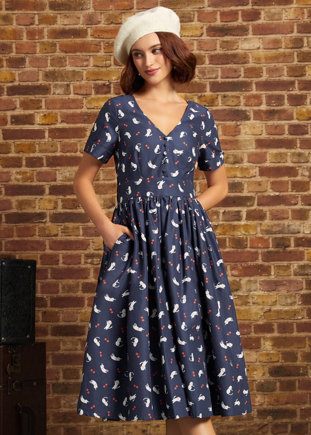 A woman wearing a navy blue ModCloth short-sleeve dress with a white and red cat print, paired with a cream beret, standing against a rustic brick wall.