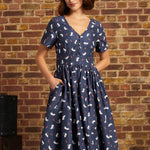 A woman wearing a navy blue ModCloth short-sleeve dress with a white and red cat print, paired with a cream beret, standing against a rustic brick wall.
