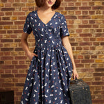 A woman wearing a navy blue short-sleeve midi dress with a white and red cat print, a cream beret, and holding a vintage suitcase against a brick wall background.