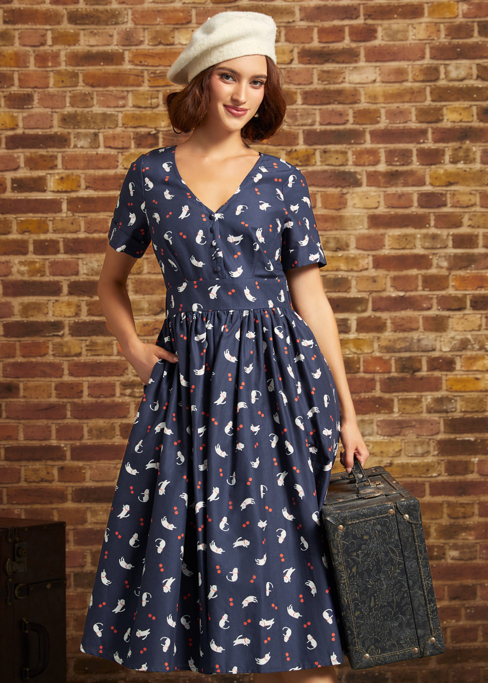 A woman wearing a navy blue short-sleeve midi dress with a white and red cat print, a cream beret, and holding a vintage suitcase against a brick wall background.