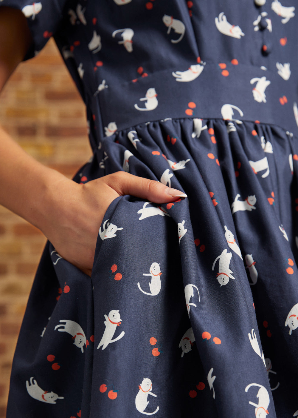 Close-up of a hand tucked into the side pocket of a navy cat print fit-and-flare dress, showing the gathered waist and red berry motifs.