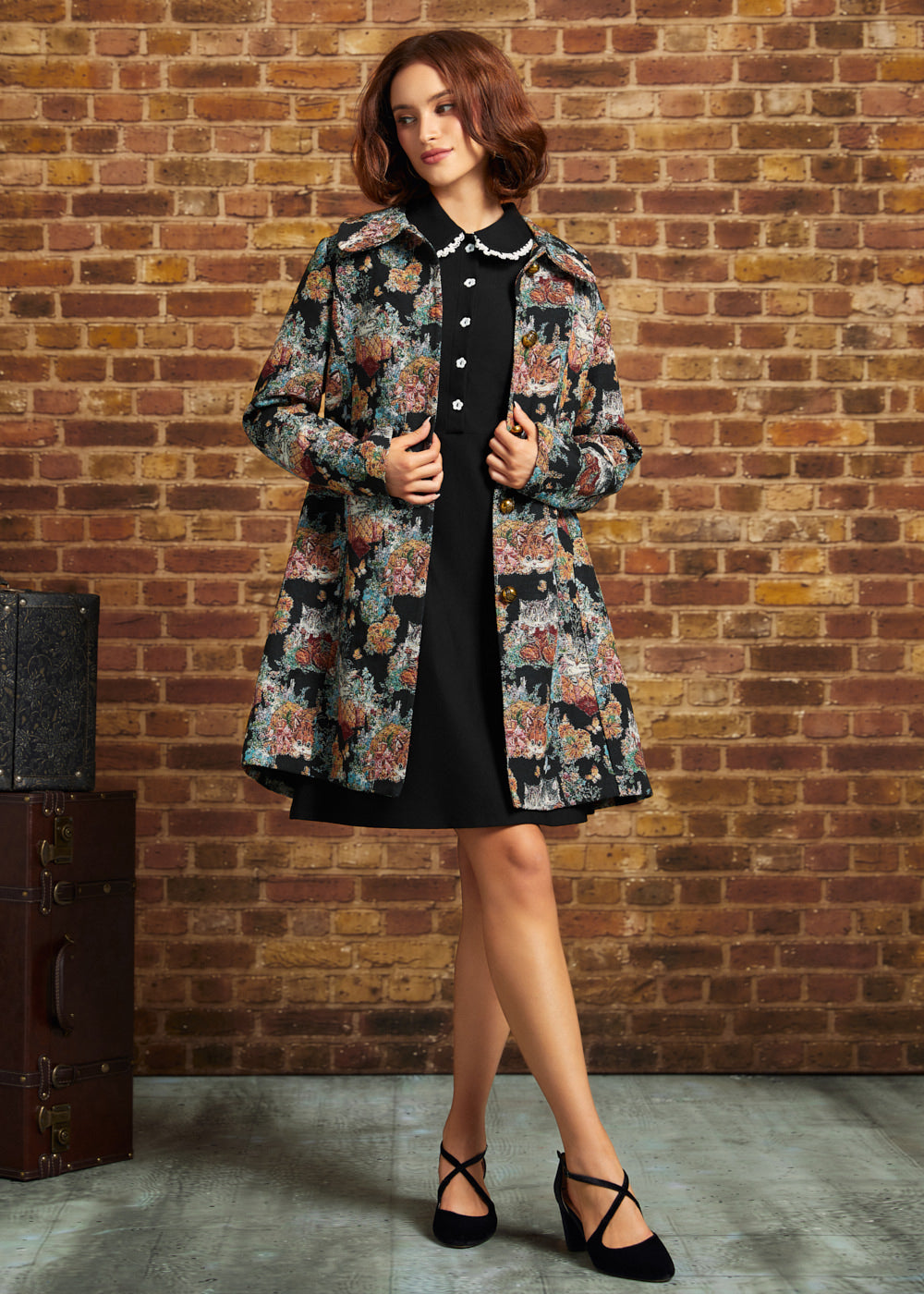 A woman stands in front of a brick wall wearing a black dress with a floral patterned coat, and black strappy heels. A vintage trunk is visible on the left side of the scene.