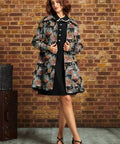 A woman stands in front of a brick wall wearing a black dress with a floral patterned coat, and black strappy heels. A vintage trunk is visible on the left side of the scene.