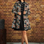 A woman stands in front of a brick wall wearing a black dress with a floral patterned coat, and black strappy heels. A vintage trunk is visible on the left side of the scene.