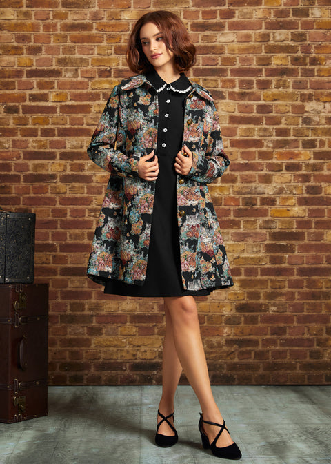 A woman stands in front of a brick wall wearing a black dress with a floral patterned coat, and black strappy heels. A vintage trunk is visible on the left side of the scene.
