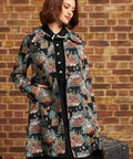 A woman models a black longline coat with a colorful floral and animal print, worn over a black dress, standing against a brick wall holding a vintage suitcase.