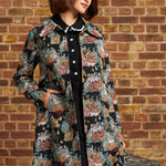 A woman models a black longline coat with a colorful floral and animal print, worn over a black dress, standing against a brick wall holding a vintage suitcase.