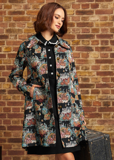 A woman models a black longline coat with a colorful floral and animal print, worn over a black dress, standing against a brick wall holding a vintage suitcase.